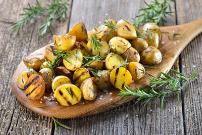 Gourmet-Rosmarin (Salvia rosmarinus) garniert gegrillte kleine Kartoffeln, die auf einem Holzbrett angerichtet sind. Die Kartoffeln sind mit frischen Rosmarinzweigen und etwas Meersalz bestreut, was dem Gericht eine aromatische und rustikale Note verleiht.