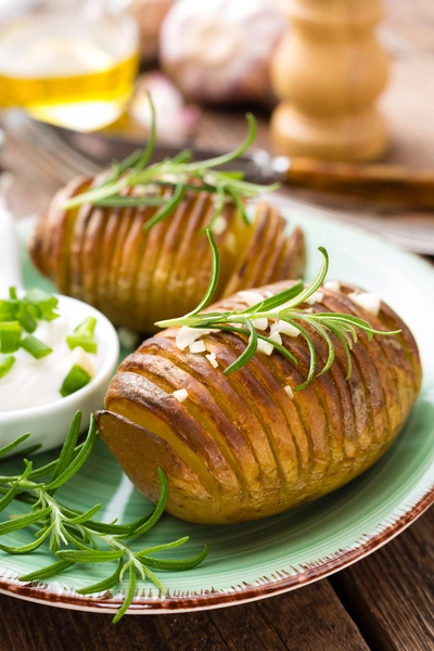 Hängender Rosmarin (Salvia rosmarinus) dekoriert gebackene Hasselback-Kartoffeln, die mit frischen Rosmarinzweigen garniert sind. Die Kartoffeln werden auf einem Teller serviert und von einer Portion Kräuterquark begleitet.