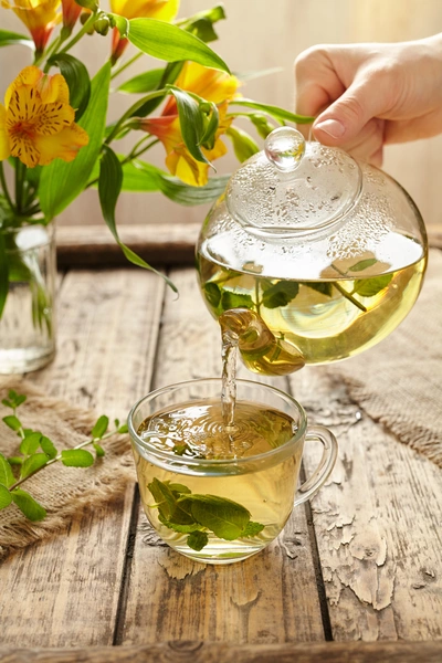 Eine Hand gießt Salbeitee aus einer gläsernen Teekanne in eine Tasse auf einem rustikalen Holztisch. Im Hintergrund steht ein Blumenstrauß mit gelben Blüten.