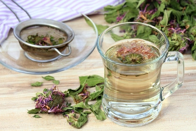 Ein Glas mit hellem Goldmelissen-Tee, in dem eine Blüte schwimmt, steht auf einem Holztisch. Daneben liegen getrocknete Blüten und Blätter, sowie ein Teesieb auf einer Glasuntertasse. Im Hintergrund ist ein lila-weiß gestreiftes Tuch zu sehen.