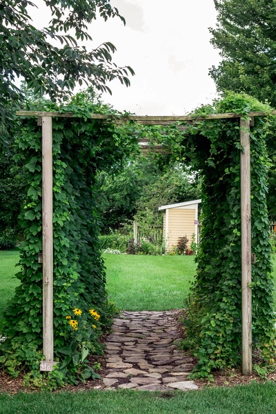 Ein mit Hopfen bewachsener Holzbogen markiert den Eingang zu einem Garten mit einem gepflasterten Weg. Im Hintergrund sind eine grüne Rasenfläche, Bäume und ein gelbes Gartenhaus zu sehen.