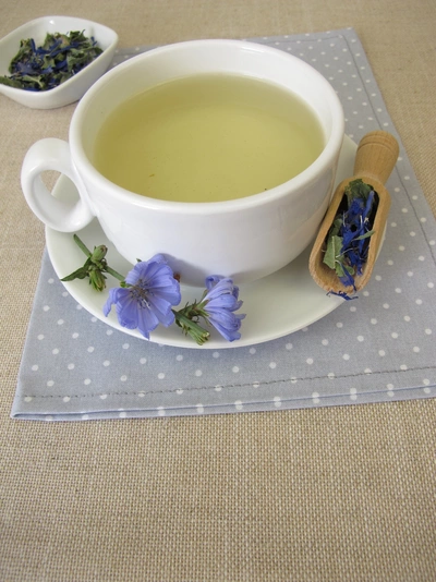 Eine weiße Tasse mit hellgelbem Wegwartentee auf einem Teller, dekoriert mit blauen Wegwartenblüten. Daneben liegt ein Holzlöffel mit getrockneten Blüten. Der Hintergrund zeigt eine beige Tischdecke mit einer grauen Serviette.