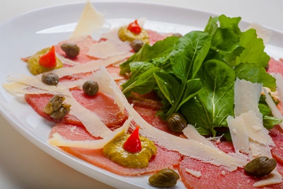 Ein Gericht aus dünn geschnittenem Rindfleisch, garniert mit Wasabi-Rucola, Parmesanflocken und Kapern. Rote Chilipaste und grüne Blätter ergänzen das kulinarische Arrangement auf einem weißen Teller.