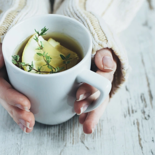 Eine Person in einem cremefarbenen Strickpullover hält eine weiße Tasse mit heißem Tee, garniert mit Zitronenscheiben und echtem Thymian, auf einem rustikalen Holztisch.