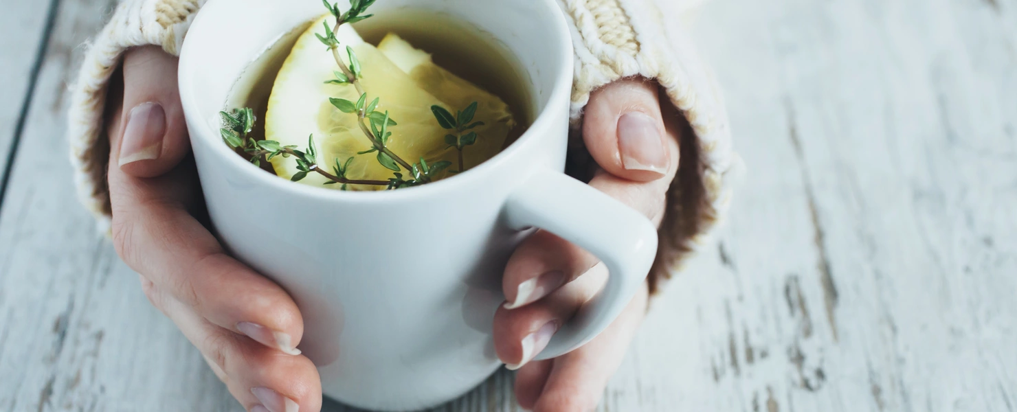 Eine Person in einem cremefarbenen Strickpullover hält eine weiße Tasse mit heißem Tee, garniert mit Zitronenscheiben und echtem Thymian, auf einem rustikalen Holztisch.