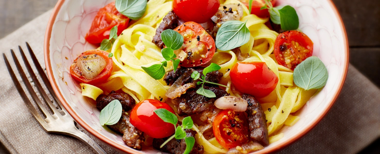 Eine Schüssel Pasta mit Tomaten, Kräutern und Oliven liegt auf einem rustikalen Holztisch. Daneben befindet sich eine Stoffserviette mit einer Gabel, während frische Oreganoblätter die Speise garnieren.
