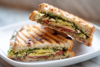 Zwei gegrillte Sandwich-Hälften auf einem weißen Teller, gefüllt mit Rucola, Tomaten, Artischocken und geschmolzenem Käse. Das Brot hat goldbraune Grillstreifen. Eine Hälfte lehnt an der anderen, die Füllung ist sichtbar. Unscharfer Hintergrund.