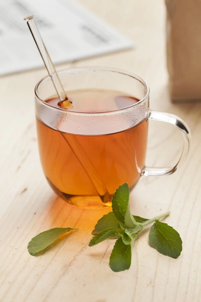 Ein Glas Tee, vermutlich mit Süßkraut (Stevia rebaudiana), wird auf einem Holztisch serviert. Ein frisches Blatt Süßkraut liegt daneben, und ein Tee-Rührstäbchen befindet sich im Glas. Der Tee hat eine goldene Farbe.