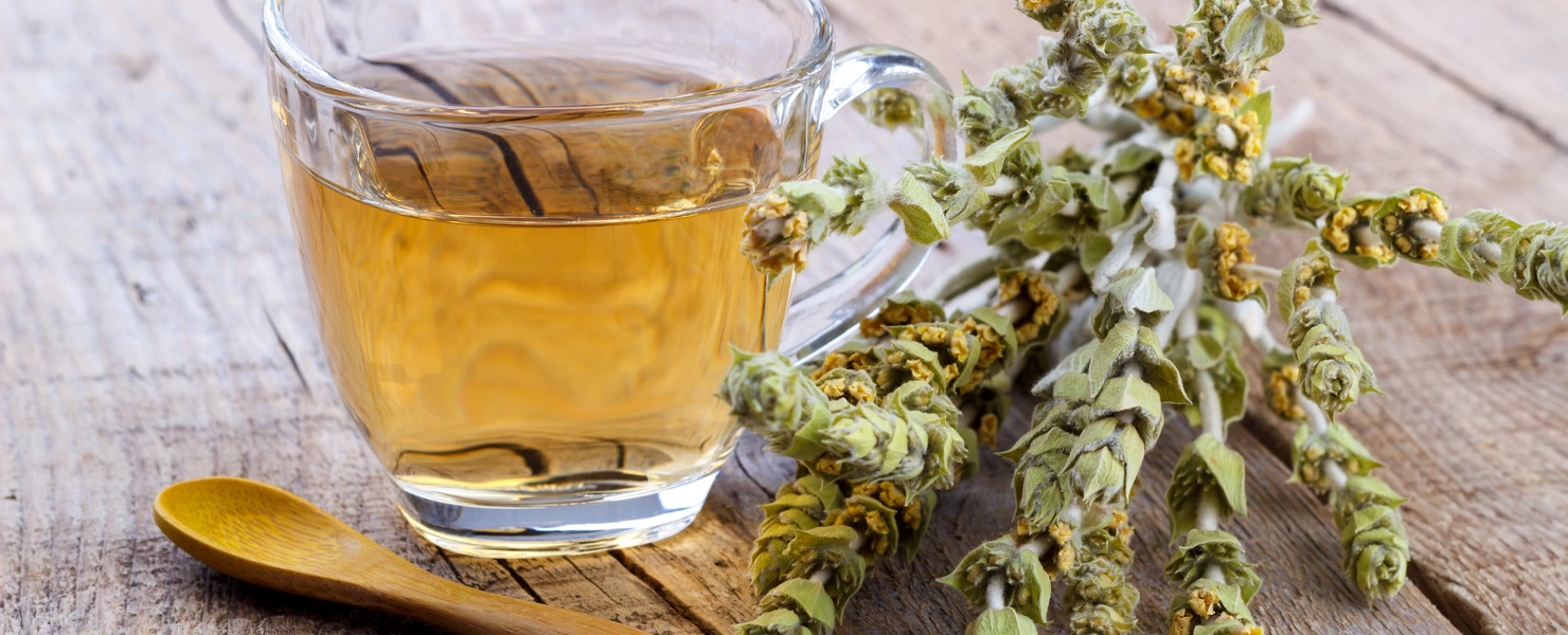 Ein Glas mit goldfarbenem griechischem Bergtee auf einer rustikalen Holzoberfläche. Daneben liegen getrocknete Bergtee-Zweige mit kleinen gelben Blüten und ein Holzlöffel.