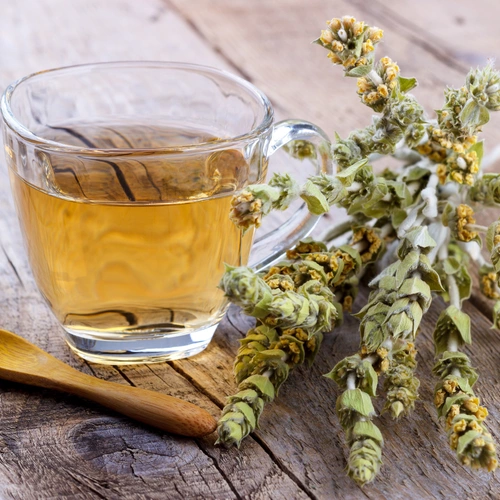 Ein Glas mit goldfarbenem griechischem Bergtee auf einer rustikalen Holzoberfläche. Daneben liegen getrocknete Bergtee-Zweige mit kleinen gelben Blüten und ein Holzlöffel.