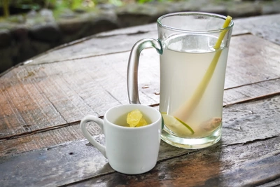 Glaskrug mit Zitronengras-Tee, in dem ein frischer Zitronengrasstängel schwimmt, daneben eine kleine weiße Tasse mit einer Zitronenscheibe. Beides steht auf einem rustikalen Holztisch mit sichtbarer Maserung, im Hintergrund eine Natursteinmauer.