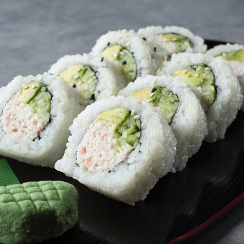 Ein Teller mit frischen Sushi-Rollen, gefüllt mit Avocado und Krabben, wird serviert. Neben den Rollen liegen Wasabi und eingelegter Ingwer, was das Gericht ergänzt.