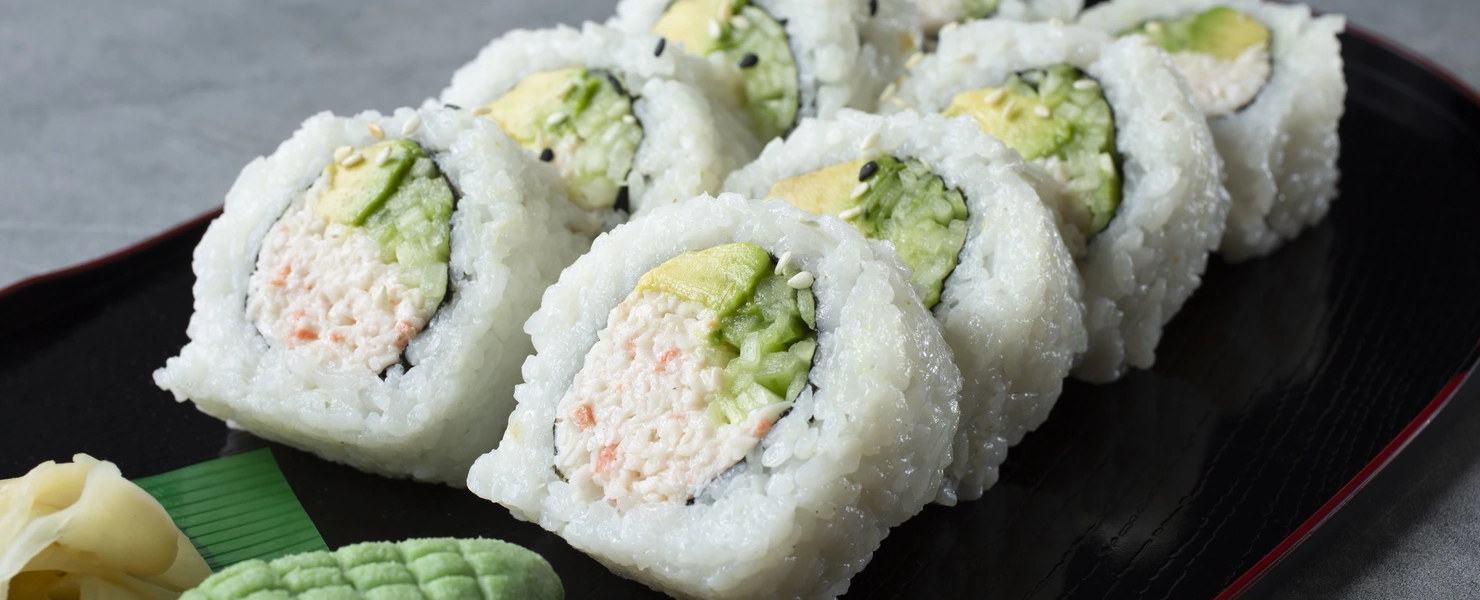 Ein Teller mit frischen Sushi-Rollen, gefüllt mit Avocado und Krabben, wird serviert. Neben den Rollen liegen Wasabi und eingelegter Ingwer, was das Gericht ergänzt.