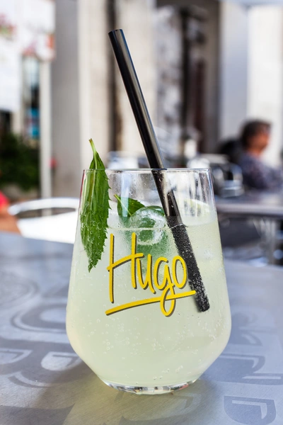 Ein Glas Hugo-Cocktail mit frischer Hugo-Minze und Strohhalm auf einem Tisch im Außenbereich, im Hintergrund unscharf Menschen und Gebäude.