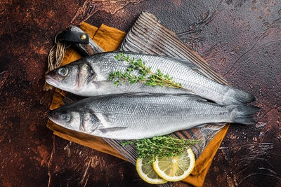 Zwei frische Fische liegen auf einem Holzbrett mit Zitronen-Thymian-Zweigen und Zitronenscheiben. Die dunkle, rustikale Unterlage verstärkt den Kontrast der Zutaten.