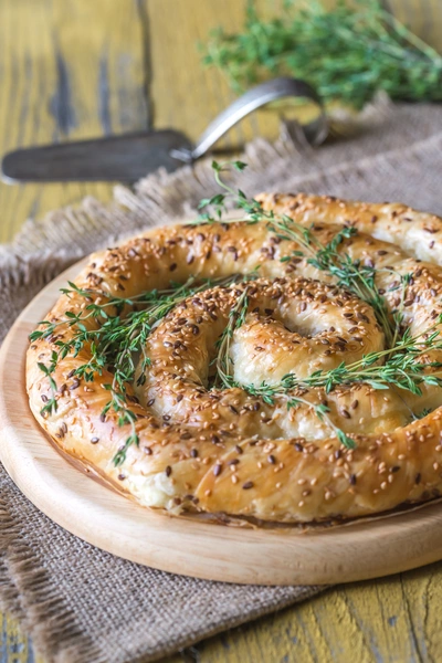 Eine goldbraune, spiralförmige Blätterteig-Pastete mit Sesam liegt auf einem runden Holzbrett, garniert mit frischem Orangen-Thymian. Im Hintergrund liegt ein Messer auf einer rustikalen Stoffunterlage.