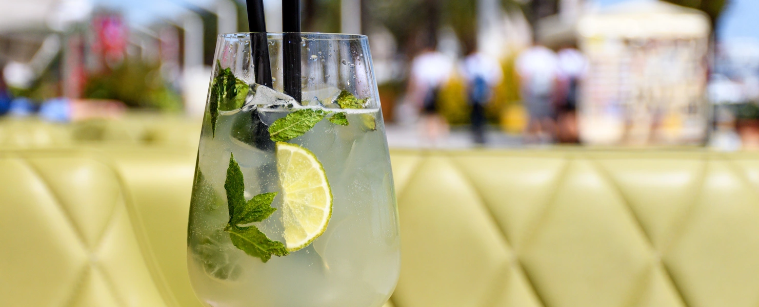 Ein Glas Hugo mit Minzblättern, Limettenscheiben und zwei schwarzen Strohhalmen, serviert auf einem gelben Polsterhintergrund. Im verschwommenen Hintergrund sind Palmen, Menschen und eine sommerliche Strandpromenade zu erkennen.