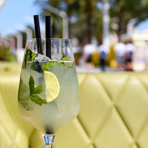 Ein Glas Hugo mit Minzblättern, Limettenscheiben und zwei schwarzen Strohhalmen, serviert auf einem gelben Polsterhintergrund. Im verschwommenen Hintergrund sind Palmen, Menschen und eine sommerliche Strandpromenade zu erkennen.