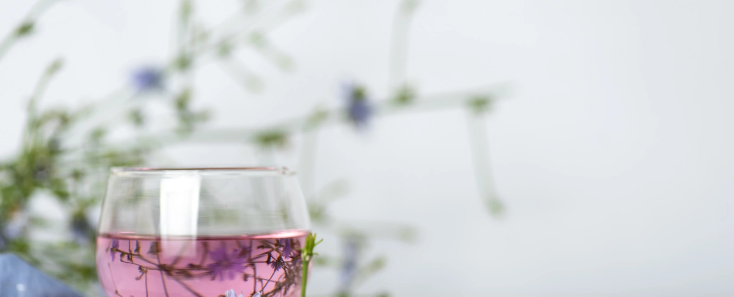 Ein Glas mit rosafarbenem Wegwartentee auf einer kleinen Holzscheibe, dekoriert mit einer blauen Wegwartenblüte. Im Hintergrund sind unscharfe Wegwartenstängel mit weiteren Blüten sichtbar.