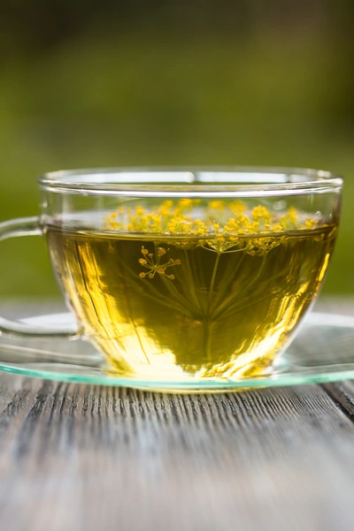Eine Tasse mit goldenem Fencheltee auf einer Holzoberfläche. In der transparenten Glastasse schwimmen kleine gelbe Fenchelblüten. Der Hintergrund ist unscharf und grünlich.