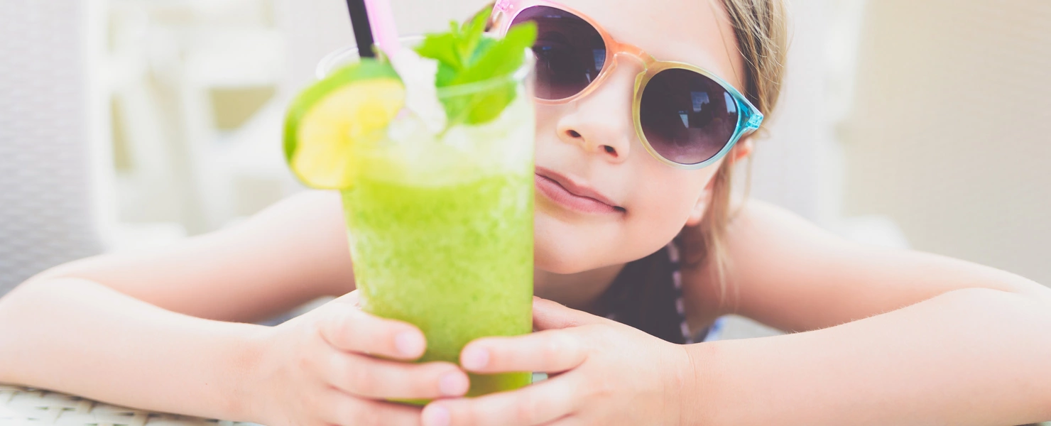 Ein Kind mit Sonnenbrille hält ein Glas grünen Smoothie, dekoriert mit Minzblättern, einer Zitronenscheibe und einem Strohhalm. Es sitzt an einem Tisch in einer sonnigen Umgebung.