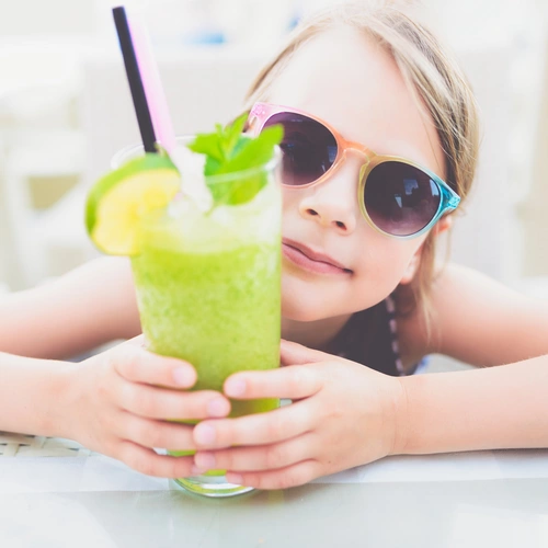 Ein Kind mit Sonnenbrille hält ein Glas grünen Smoothie, dekoriert mit Minzblättern, einer Zitronenscheibe und einem Strohhalm. Es sitzt an einem Tisch in einer sonnigen Umgebung.