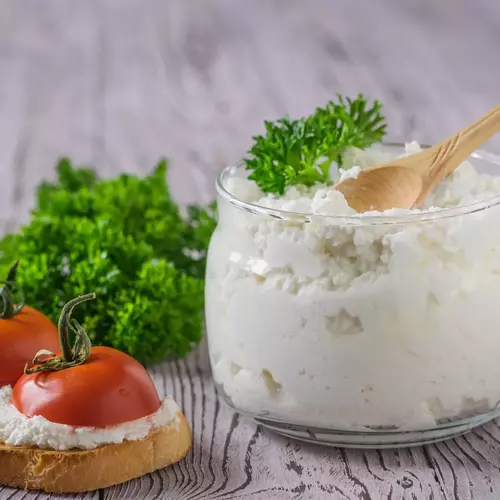 Ein Glas mit frischem Quark, garniert mit frischer Krauser Petersilie (Petroselinum crispum), neben einer belegten Brotscheibe mit Tomaten. Die Zutaten sind auf einem rustikalen Holzuntergrund angerichtet und verleihen dem Bild eine einfache, hausgemachte Atmosphäre.