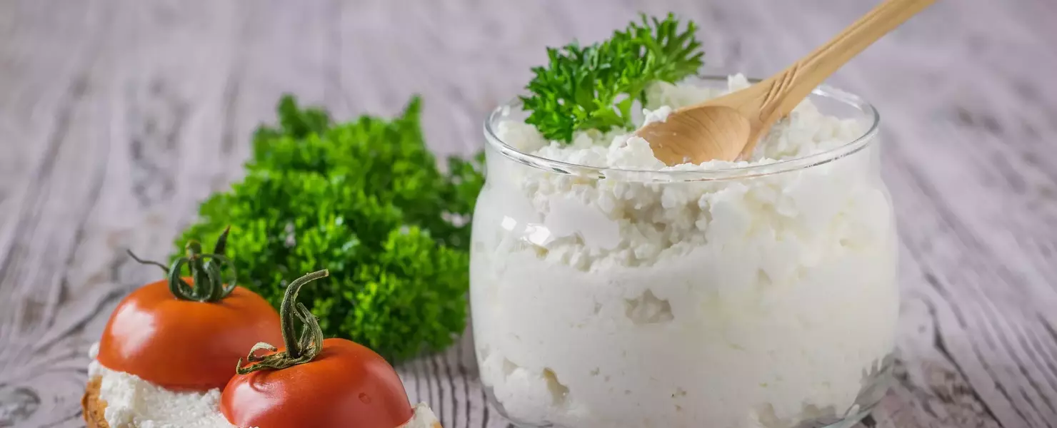 Ein Glas mit frischem Quark, garniert mit frischer Krauser Petersilie (Petroselinum crispum), neben einer belegten Brotscheibe mit Tomaten. Die Zutaten sind auf einem rustikalen Holzuntergrund angerichtet und verleihen dem Bild eine einfache, hausgemachte Atmosphäre.