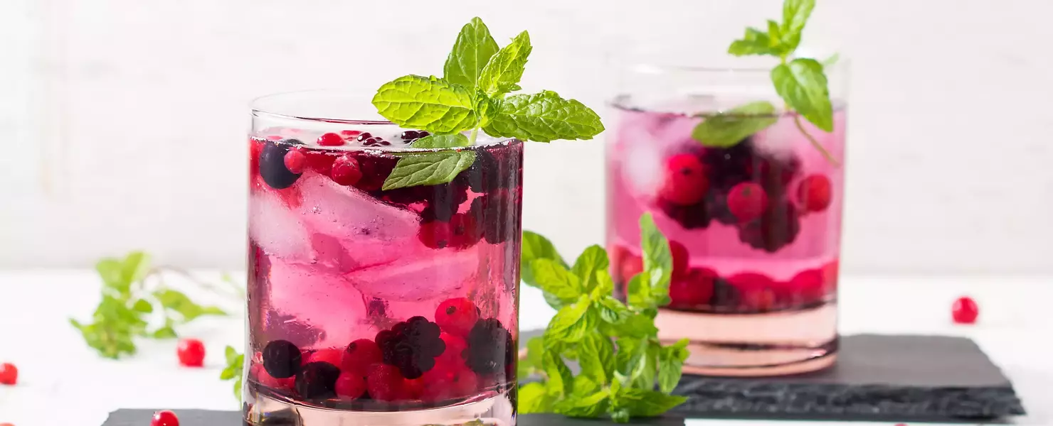 Zwei Gläser Johannisbeer-Minze-Getränk mit frischen Johannisbeeren, Minzblättern und Eiswürfeln auf dunklen Schieferplatten. Im Hintergrund helle Wand und verstreute Beeren als Dekoration.