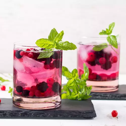 Zwei Gläser Johannisbeer-Minze-Getränk mit frischen Johannisbeeren, Minzblättern und Eiswürfeln auf dunklen Schieferplatten. Im Hintergrund helle Wand und verstreute Beeren als Dekoration.