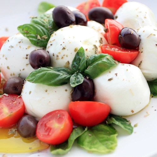 Ein Teller mit einem klassischen italienischen Gericht, bestehend aus frischen Mozzarella-Kugeln, saftigen, halbierten Kirschtomaten, schwarzen Oliven und aromatischen Basilikumblättern. Das Ganze ist mit etwas Olivenöl beträufelt und mit Kräutern bestreut.