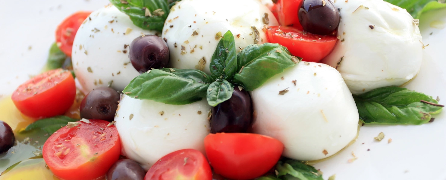 Ein Teller mit einem klassischen italienischen Gericht, bestehend aus frischen Mozzarella-Kugeln, saftigen, halbierten Kirschtomaten, schwarzen Oliven und aromatischen Basilikumblättern. Das Ganze ist mit etwas Olivenöl beträufelt und mit Kräutern bestreut.