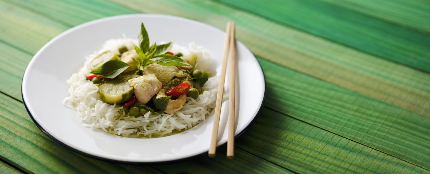 Ein Teller mit thailändischem Curry, serviert auf Reisnudeln, garniert mit Thai-Basilikum (Ocimum basilicum var. thyrsiflora) Blättern und roten Chilischoten. Die frischen Kräuter und Gewürze verleihen dem Gericht eine aromatische Note.
