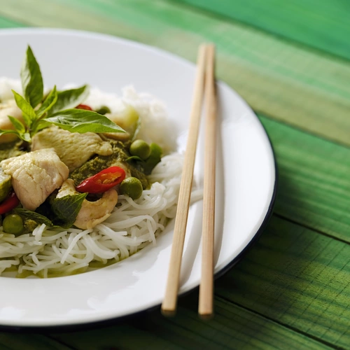 Ein Teller mit thailändischem Curry, serviert auf Reisnudeln, garniert mit Thai-Basilikum (Ocimum basilicum var. thyrsiflora) Blättern und roten Chilischoten. Die frischen Kräuter und Gewürze verleihen dem Gericht eine aromatische Note.