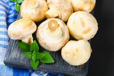Frische Champignons auf einem schwarzen Holzbrett, zusammen mit frischen grünen Kräutern, auf einem blauen karierten Tuch ausgebreitet.
