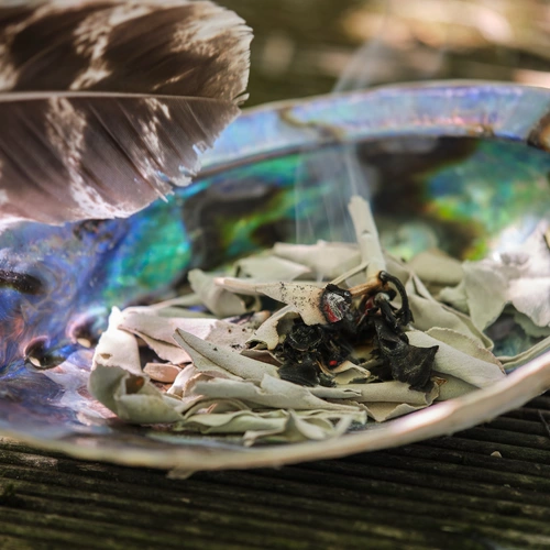 Eine schillernde Muschelschale mit glühenden Räuchersalbei-Blättern, aus denen Rauch aufsteigt, liegt auf einer Holzunterlage, daneben eine große Feder.
