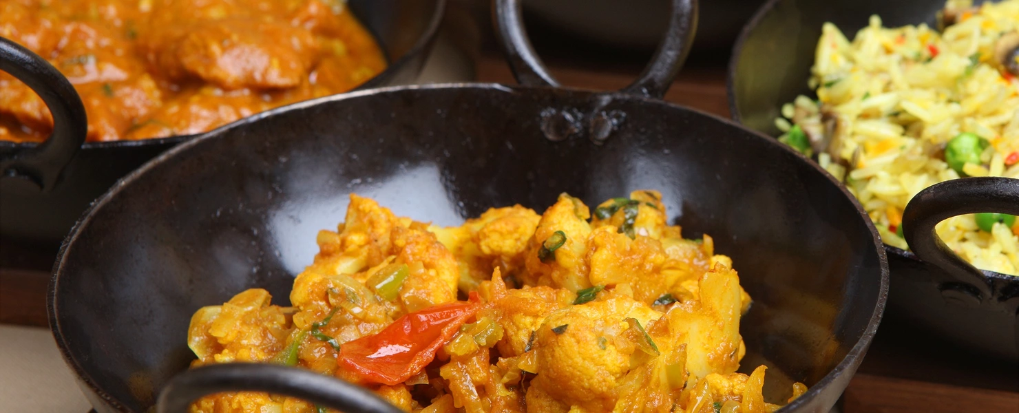 Currykraut (Murraya koenigii) in einer Reihe von schwarzen Schalen mit verschiedenen Currygerichten, darunter ein gelbes Curry mit Gemüse, das frisch serviert wird. Einige Schalen enthalten auch eine Reisbeilage.