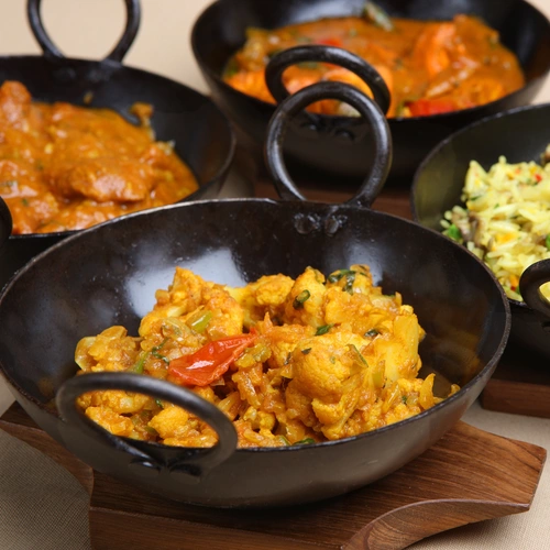 Currykraut (Murraya koenigii) in einer Reihe von schwarzen Schalen mit verschiedenen Currygerichten, darunter ein gelbes Curry mit Gemüse, das frisch serviert wird. Einige Schalen enthalten auch eine Reisbeilage.