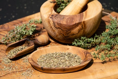 Bergbohnenkraut (Satureja montana) ist in einem Mörser aus Holz zerkleinert worden. Die Kräuter, frisch und getrocknet, liegen auf einem Holzbrett. Der Mörser und das Zerkleinern der Blätter deuten auf die Zubereitung von Gewürzen für die Küche hin.
