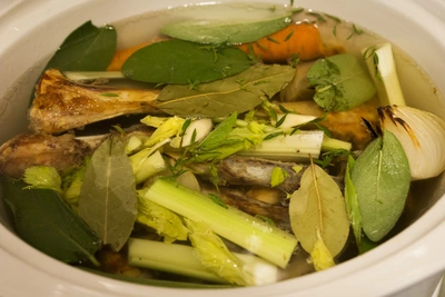 Ein Topf mit einer klaren Brühe, in der verschiedene Gemüse wie Karotten, Lauch und Sellerie sowie Lorbeerblätter schwimmen. Die Zutaten sind gut sichtbar und frisch, was auf eine aromatische Suppe hinweist.