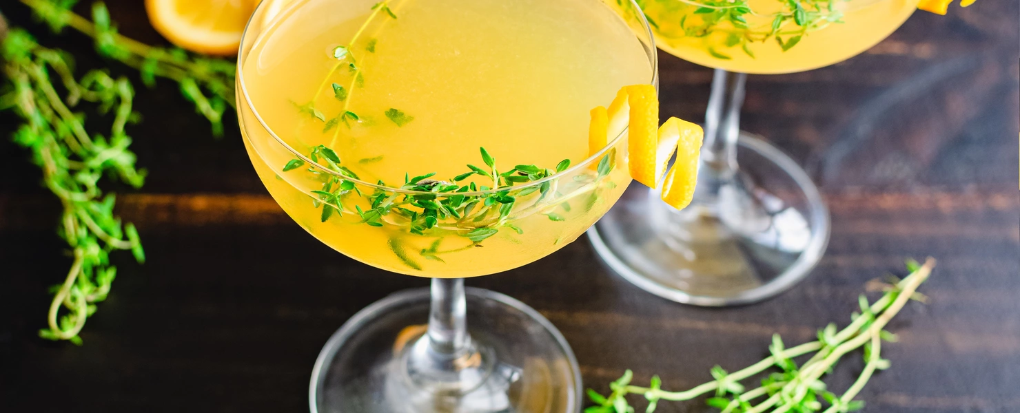 Zwei Cocktails mit gelber Flüssigkeit in eleganten Gläsern, garniert mit Zitronen-Thymian-Zweigen und Zitronenzesten. Im Hintergrund liegen halbierte Zitronen auf einer dunklen Oberfläche.