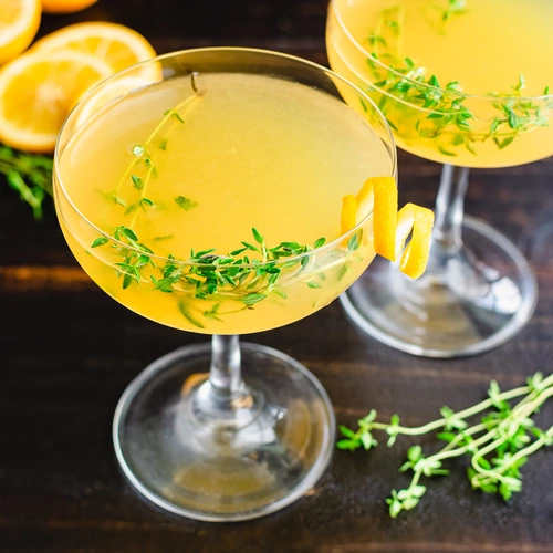 Zwei Cocktails mit gelber Flüssigkeit in eleganten Gläsern, garniert mit Zitronen-Thymian-Zweigen und Zitronenzesten. Im Hintergrund liegen halbierte Zitronen auf einer dunklen Oberfläche.