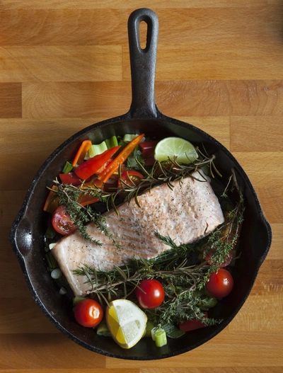 Ein Stück gebratener Fisch liegt in einer schwarzen Pfanne, umgeben von frischem Thymian, Tomaten, Paprika und Zitronenscheiben. Die Pfanne steht auf einem Holztisch und zeigt eine farbenfrohe, gesunde Mahlzeit.