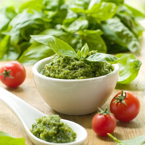 Das Bild zeigt eine Schale mit grünem Pesto, dekoriert mit frischen Basilikumblättern. Im Hintergrund sind üppige Blätter des Berg-Basilikums (Ocimum basilicum 'Mountain') zu sehen. Daneben liegen kleine, rote Tomaten und ein Löffel mit etwas Pesto, was das frische Aroma des Gerichts hervorhebt.