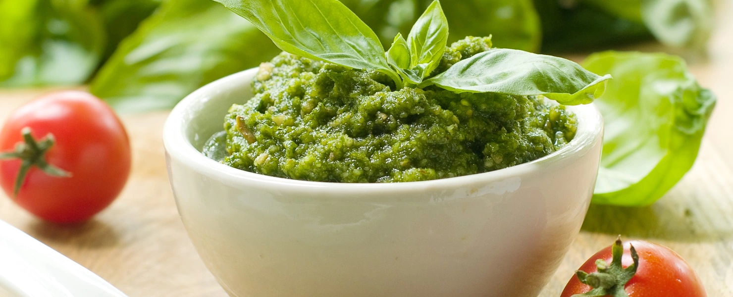 Das Bild zeigt eine Schale mit grünem Pesto, dekoriert mit frischen Basilikumblättern. Im Hintergrund sind üppige Blätter des Berg-Basilikums (Ocimum basilicum 'Mountain') zu sehen. Daneben liegen kleine, rote Tomaten und ein Löffel mit etwas Pesto, was das frische Aroma des Gerichts hervorhebt.