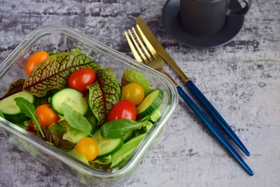 Ein Salat mit frischen Tomaten, Gurken und Blättern von Blut-Sauerampfer (Rumex sanguineus). Der Salat ist in einem Glasbehälter angerichtet, umgeben von glänzendem Besteck und einer Tasse, was das Bild zu einer einladenden und stilvollen Mahlzeit macht.