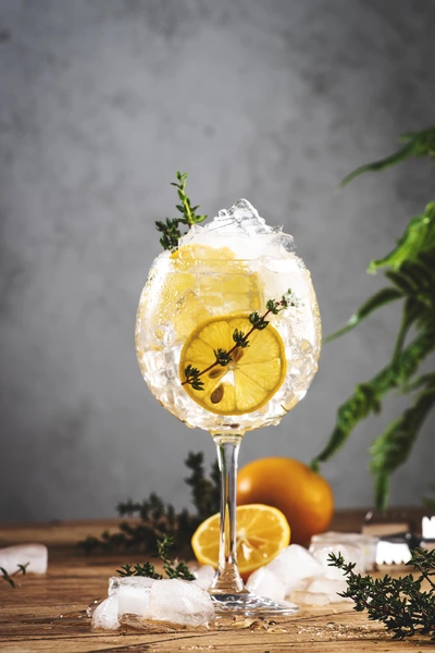 Ein erfrischendes Getränk in einem Weinglas mit Eiswürfeln, einer Zitronenscheibe und einem Zweig Orangen-Thymian, auf einem rustikalen Holztisch mit frischen Kräutern und Orangen im Hintergrund.