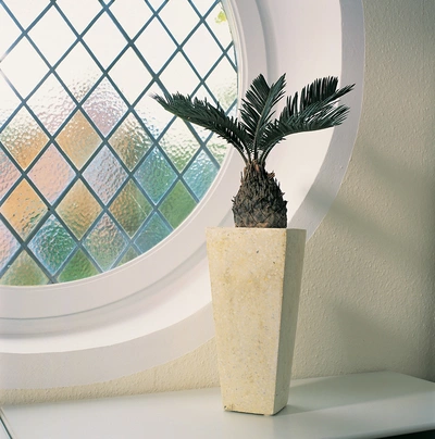 Ein Palmfarn in einem hohen, cremefarbenen Topf, platziert auf einem Tisch vor einem runden Fenster mit geometrischem Muster