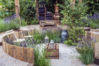 Ein kreisförmiger Garten mit Lavendel, umrandet von niedrigen Holzbegrenzungen. In der Mitte stehen Holzkisten mit Laternen. Im Hintergrund befinden sich Kupfergefäße, gestapelte Lavendelsträuße und eine Rankpflanze an einem Holzpfosten.