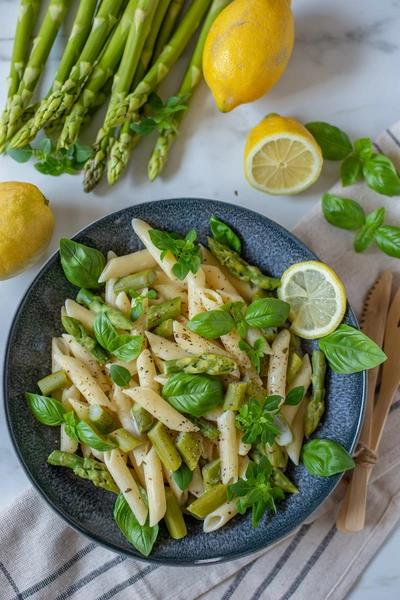  Ein Teller mit Penne, grünem Spargel und italienischem Basilikum (Ocimum basilicum). Der Teller ist dekoriert mit frischen Basilikumblättern und Zitronenscheiben. Im Hintergrund liegen frische Spargelstangen und Zitronen auf einem hellen Untergrund.